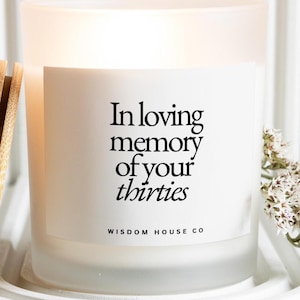 May include: A frosted glass candle with the text "In loving memory of your thirties" in black. The candle is on a white tray with a wooden object to the left and white flowers to the right. The brand name "Wisdom House Co" is at the bottom.