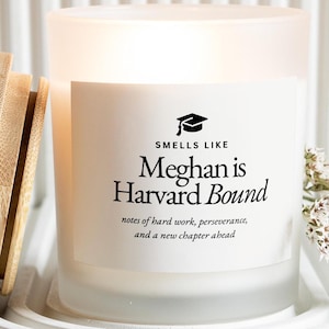 May include: A frosted glass candle with a wooden lid, resting on a white, textured surface. The candle has a label that reads "Smells Like Meghan is Harvard Bound" with additional text below. A graduation cap graphic is also present.