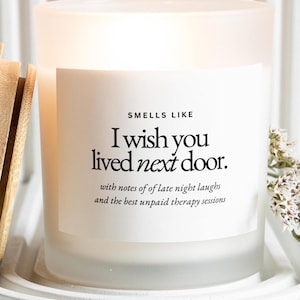 May include: A white scented candle in a frosted glass jar. The candle has a label with the text "I wish you lived next door." The candle is on a white tray with a bamboo item and white flowers.