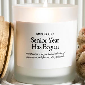 May include: A lit candle in a frosted glass jar with a bamboo lid. The white label features black text reading: SMELLS LIKE Senior Year Has Begun, notes of last-first days, a packed calendar of countdowns, and finally ruling the school.