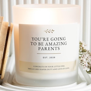 Op de afbeelding: Een matglazen kaars met een houten deksel, met de tekst "YOU'RE GOING TO BE AMAZING PARENTS" en "EST. 2026". De kaars heeft een wit etiket met een bloemenontwerp en een boodschap over een pasgeborene. Een takje witte bloemen ligt naast de kaars.