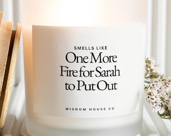 May include: A frosted glass candle with a wooden lid. The candle has a white label with the text "Smells Like One More Fire for Sarah to Put Out" and "Wisdom House Co." The candle is on a white tray.