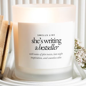 Könnte beinhalten: Eine weiße Kerze aus mattiertem Glas mit Holzdeckel, auf einem weißen Tablett. Die Kerze hat ein Etikett mit der Aufschrift "Smells Like she's writing a bestseller" mit zusätzlichem Text. Ein Zweig weißer Blumen befindet sich neben der Kerze.