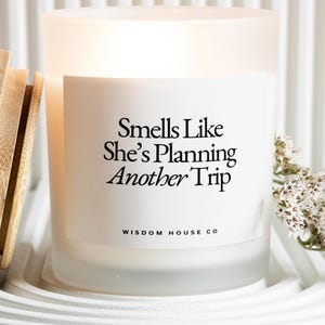 May include: Frosted glass candle with the text "Smells Like She's Planning Another Trip" and "Wisdom House Co." printed on a white label. The candle has a wooden lid and sits on a white, textured surface. Dried flowers are to the right.