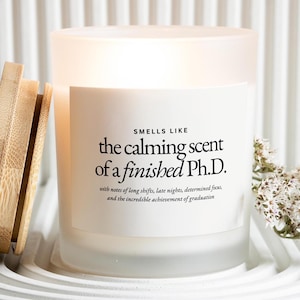 May include: A frosted glass candle with a wooden lid, labeled "Smells Like the calming scent of a finished Ph.D." The candle sits on a white tray with a floral arrangement. The text on the label describes the scent.