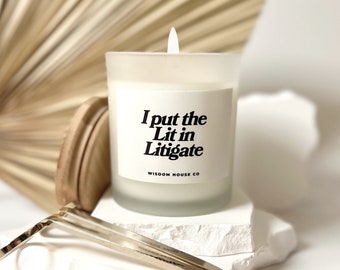 May include: A white candle with a frosted glass jar and a black label that reads "I put the Lit in Litigate" with the text "Wisdom House Co" below. The candle is sitting on a white rock with a gold metal candle snuffer.