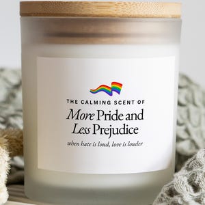 May include: A frosted glass candle jar with a light wood lid. The white label features a rainbow flag icon and black text reading: THE CALMING SCENT OF More Pride and Less Prejudice when hate is loud, love is louder. The jar is cylindrical.