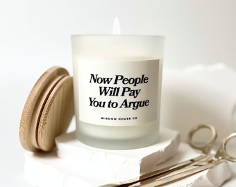 May include: A white frosted glass candle jar with a black label that reads "Now People Will Pay You to Argue" and "Wisdom House Co.". The candle is on a white surface with a wooden lid and a gold candle snuffer.