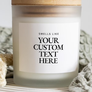 May include: A frosted glass candle jar with a light wood lid. The white rectangular label on the front features black text that reads: SMELLS LIKE YOUR CUSTOM TEXT HERE. The candle container has a cylindrical shape and a smooth, matte finish.