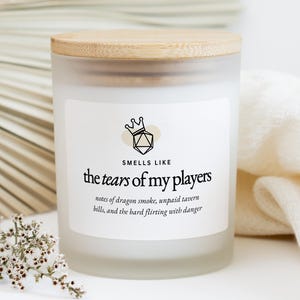 Könnte beinhalten: Ein Kerzenglas aus mattiertem Glas mit einem Holzdeckel. Auf dem Etikett steht "Smells Like the Tears of My Players" mit Begleittext. Die Kerze ist von einem weißen Tuch und einem Zweig mit kleinen weißen Blüten umgeben.