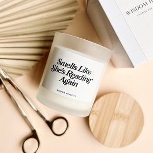 May include: A white frosted glass candle with a black label that reads "Smells Like She's Reading Again" and the text "Wisdom House Co.". The candle is sitting on a light pink surface with a wooden lid and a fan made of natural material.