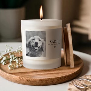 May include: A lit white candle with a photo of a dog and the name "Sadie" on the label. The candle sits on a wooden tray with a wooden lid and a sprig of white flowers.