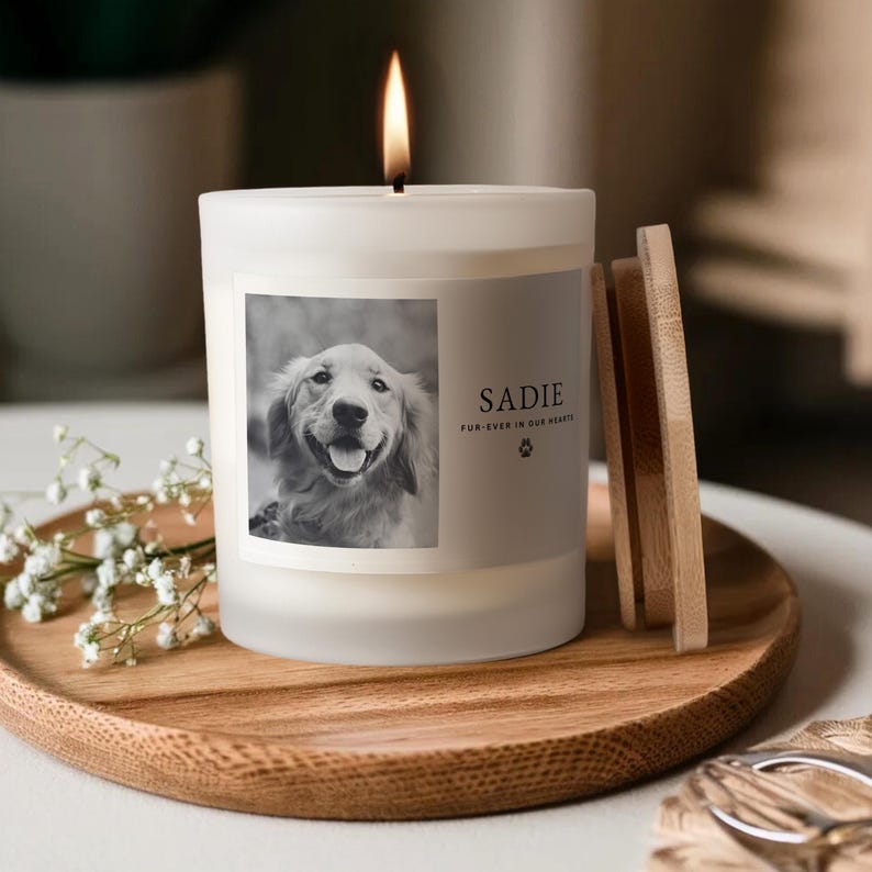 May include: A lit white candle with a photo of a dog and the name "Sadie" on the label. The candle sits on a wooden tray with a wooden lid and a sprig of white flowers.