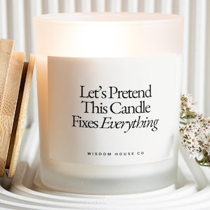 Op de afbeelding: Een witte kaars van matglas met de tekst "Let's Pretend This Candle Fixes Everything" in het zwart. De kaars staat in een witte schaal met een houten deksel en een takje witte bloemen.