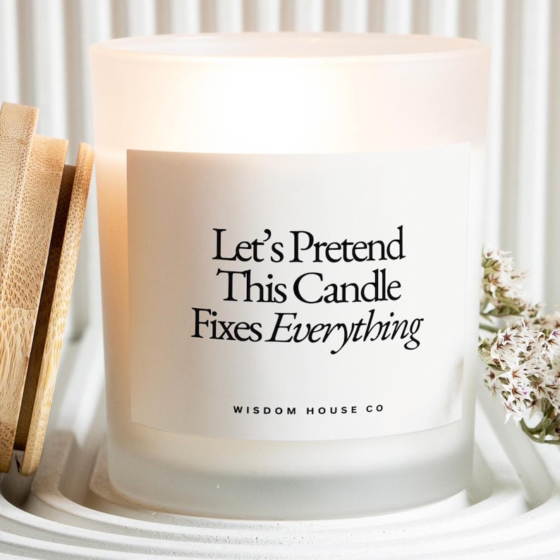 May include: A white frosted glass candle with the text "Let's Pretend This Candle Fixes Everything" in black. The candle is in a white tray with a wooden lid and a sprig of white flowers.