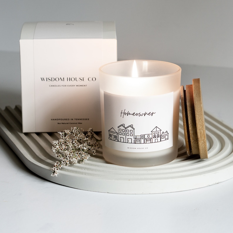 Op de afbeelding: Een witte kaars in een matglazen pot met een houten deksel. De kaars is gelabeld met "Homeowner" met een zwart-witte illustratie van huizen. De kaars staat op een wit, gebogen oppervlak met een klein boeket witte bloemen.