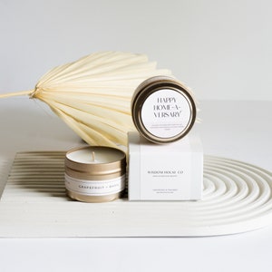 May include: Two gold candle tins with lids. The smaller tin is labeled "Grapefruit + Basil" and the larger tin is labeled "Happy Home-a-versary". Both tins are on a white rectangular surface with a circular pattern.