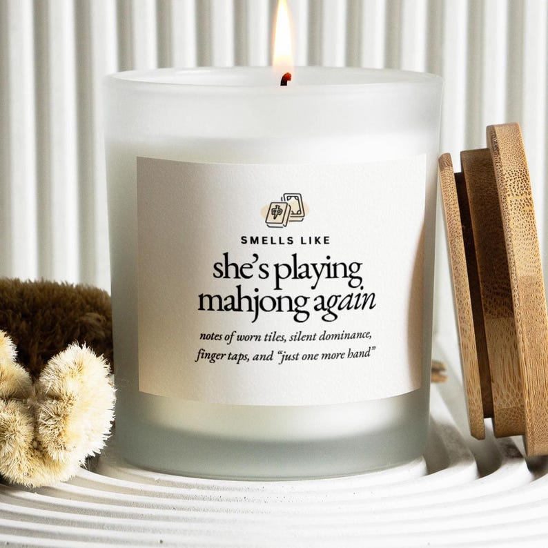 Op de afbeelding: Een brandende, matglazen kaars met een houten deksel. De kaars heeft een label met de tekst "Smells Like she's playing mahjong again". De kaars staat op een wit, geribbeld oppervlak.