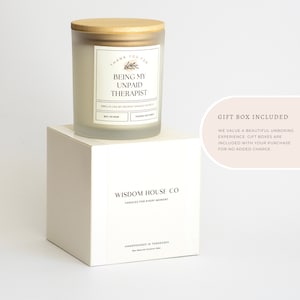 May include: White candle with a wooden lid and a label that reads "Thank you for being my unpaid therapist". The candle is on a white box with the text "Wisdom House Co. Candles for every moment" and "Handpoured in Tennessee Soy Natural Coconut Wax". The text "Gift Box Included" is in the upper right corner of the image. The text below reads "We value a beautiful unboxing experience. Gift boxes are included with your purchase for no added charge."