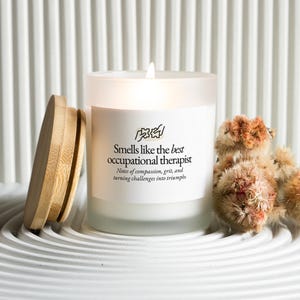 May include: A lit white candle in a frosted glass jar with a wooden lid. The candle has text that reads "Smells like the best occupational therapist." Dried, fluffy, brown flowers are next to the candle.