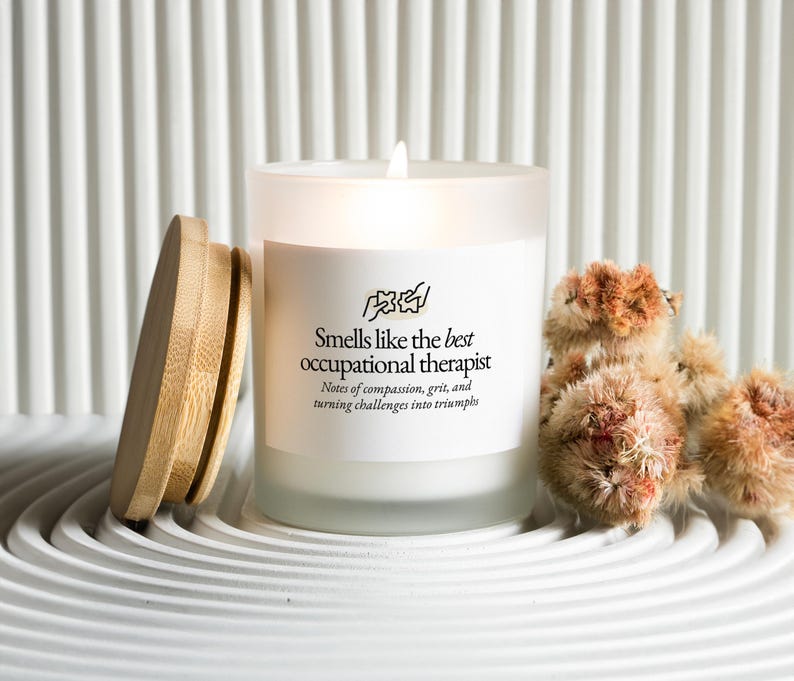 May include: A lit white candle in a frosted glass jar with a wooden lid. The candle has text that reads "Smells like the best occupational therapist." Dried, fluffy, brown flowers are next to the candle.