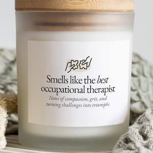 May include: A frosted glass candle with a wooden lid. The candle has a white label with the text "Smells like the best occupational therapist" and a graphic. The candle is on a striped surface.
