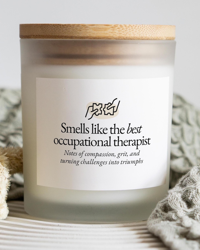 May include: A frosted glass candle with a wooden lid. The candle has a white label with the text "Smells like the best occupational therapist" and a graphic. The candle is on a striped surface.