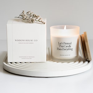 May include: A white candle in a frosted glass jar with the text "Let's Pretend This Candle Fixes Everything." A white box with the text "Wisdom House Co" and "Candles for Every Moment" is next to the candle. The candle and box are on a white, circular tray.