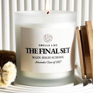 May include: A frosted glass candle with a lit wick and a wooden lid. The label features a volleyball icon and the text: SMELLS LIKE THE FINAL SET, MAIN HIGH SCHOOL, Amanda Class of 2027. The candle emits a warm glow from the flame at the top.