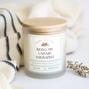 May include: A white candle with a bamboo lid and a label that reads "Thank you for being my unpaid therapist. Smells like my deepest darkest secrets. 8 oz | 45 hour. Coconut soy candle."