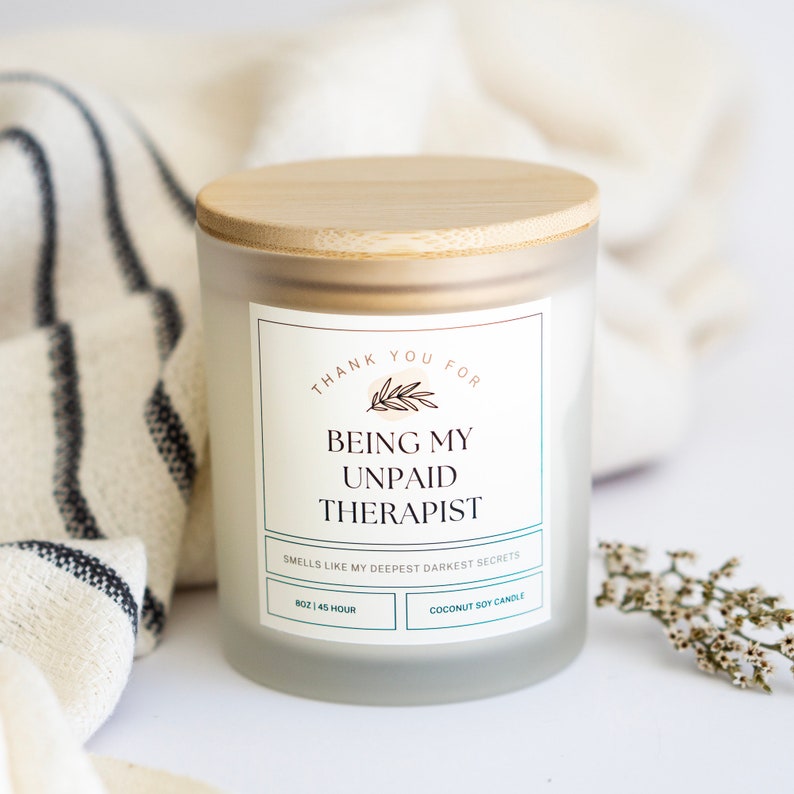 May include: A white candle with a bamboo lid and a label that reads "Thank you for being my unpaid therapist. Smells like my deepest darkest secrets. 8 oz | 45 hour. Coconut soy candle."