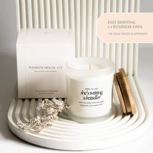 May include: A white candle in a frosted glass jar with a wooden lid. The label reads "Smells Like she's writing a bestseller." A white box with the text "Wisdom House Co" is behind the candle. The candle sits on a white, curved tray.