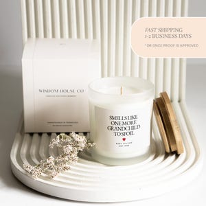 Pu&ograve; includere: Una candela bianca in un barattolo di vetro smerigliato con coperchio in legno e una scatola bianca. La candela ha il testo "Smells Like One More Grandchild To Spoil". La scatola recita "Wisdom House Co."