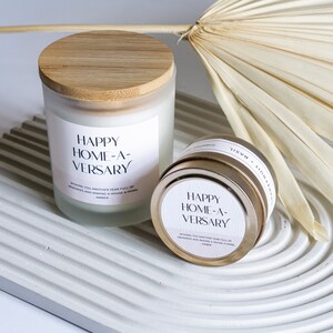 May include: Two candles with a white label that reads "Happy Home-A-Versary" and the text "Wishing you another year full of memories and making a house a home. Amber". The candles are on a white textured surface. One candle is in a glass jar with a wooden lid. The other candle is in a gold tin.