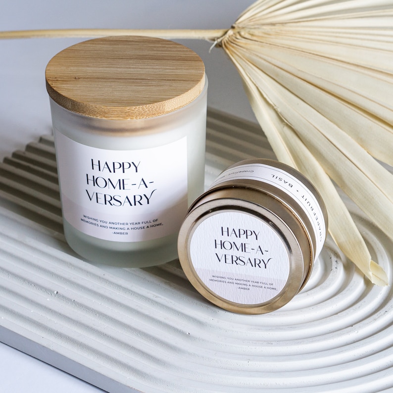 May include: Two candles with a white label that reads "Happy Home-A-Versary" and the text "Wishing you another year full of memories and making a house a home. Amber". The candles are on a white textured surface. One candle is in a glass jar with a wooden lid. The other candle is in a gold tin.