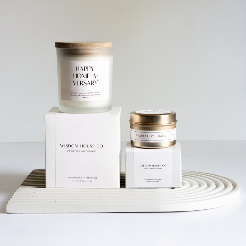 May include: Two candles, one in a glass jar with a wooden lid and the other in a metal tin. The glass jar candle has a white label with the text "HAPPY HOME-A-VERSARY". The metal tin candle has a gold label with the text "GRAPEFRUIT BASIL". Both candles are on a white platform with the text "WISDOM HOUSE CO" on the label.