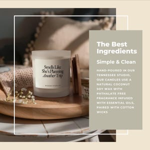 May include: A frosted glass candle with the text "Smells Like She's Planning Another Trip" on a white label. The candle sits on a wooden tray with a wooden lid and sprigs of white flowers. The background is blurred.
