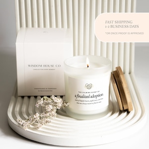 May include: A lit candle in a frosted glass jar with a wooden lid sits on a white, curved tray. The candle label reads "The Calming Scent of a finalized adoption." A white box with "Wisdom House Co" is behind the candle.