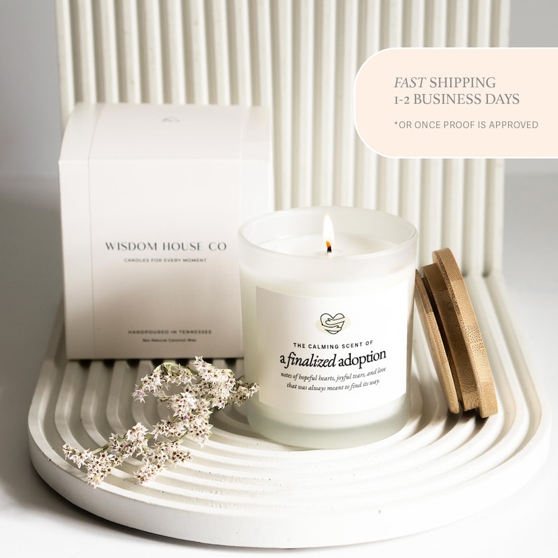 May include: A lit candle in a frosted glass jar with a wooden lid sits on a white, curved tray. The candle label reads "The Calming Scent of a finalized adoption." A white box with "Wisdom House Co" is behind the candle.