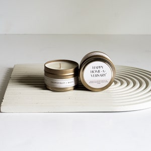 May include: Two gold candle tins with white labels. The candle on the left is labeled "Grapefruit + Basil". The candle on the right is labeled "Happy Home-a-versary".