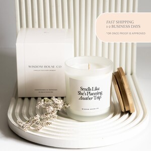 May include: A white candle in a frosted glass jar with a wooden lid, next to a white box. The candle has the text "Smells Like She's Planning Another Trip". The box reads "Wisdom House Co". A sprig of white flowers is in front.