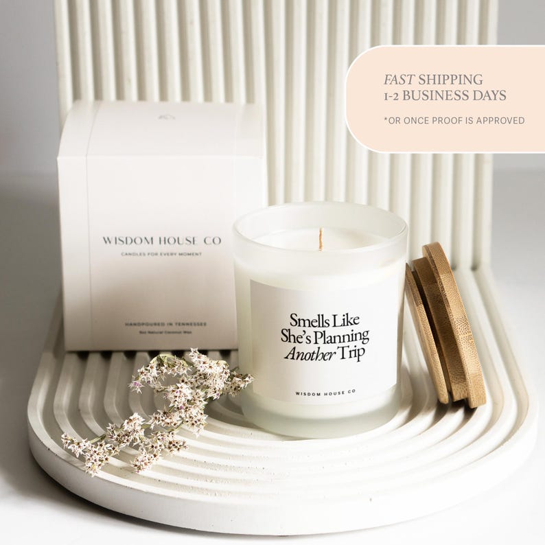 May include: A white candle in a frosted glass jar with a wooden lid, next to a white box. The candle has the text "Smells Like She's Planning Another Trip". The box reads "Wisdom House Co". A sprig of white flowers is in front.