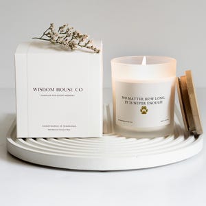 May include: A white box and a lit candle sit on a white, circular tray. The box reads "WISDOM HOUSE CO" and "CANDLES FOR EVERY MOMENT." The candle has the text "NO MATTER HOW LONG, IT IS NEVER ENOUGH." A sprig of white flowers rests on the box.