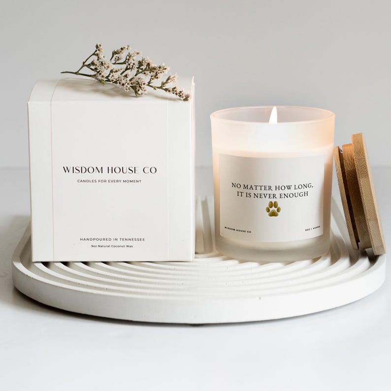 May include: A white box and a lit candle sit on a white, circular tray. The box reads "WISDOM HOUSE CO" and "CANDLES FOR EVERY MOMENT." The candle has the text "NO MATTER HOW LONG, IT IS NEVER ENOUGH." A sprig of white flowers rests on the box.