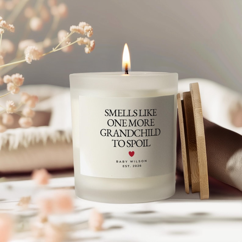 Pu&ograve; includere: Una candela bianca accesa in un barattolo di vetro smerigliato con coperchio in legno. L'etichetta recita "Smells Like One More Grandchild To Spoil" con un cuore rosso e "Baby Wilson Est. 2026". La candela &egrave; su una superficie bianca.