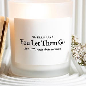 May include: A frosted glass candle jar with a white label featuring black text that reads: SMELLS LIKE You Let Them Go but still track their location. The candle is illuminated from within, casting a warm glow through the translucent glass walls.