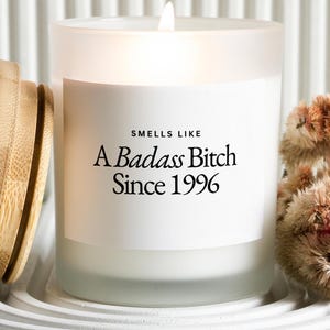 May include: A frosted glass candle in a cylindrical container with a white label featuring the black text "SMELLS LIKE A Badass Bitch Since 1996". The candle has a lit wick and is accompanied by a light-colored wooden lid placed to the left side.