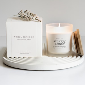 May include: A white candle in a frosted glass jar with a wooden lid, next to a white box. The candle has the text "she's writing a bestseller". The box reads "Wisdom House Co" and "Candles for every moment".