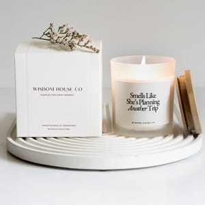 May include: A white box and a frosted glass candle with a wooden lid sit on a white, circular tray. The box reads "Wisdom House Co" and "Candles for Every Moment." The candle reads "Smells Like She's Planning Another Trip."