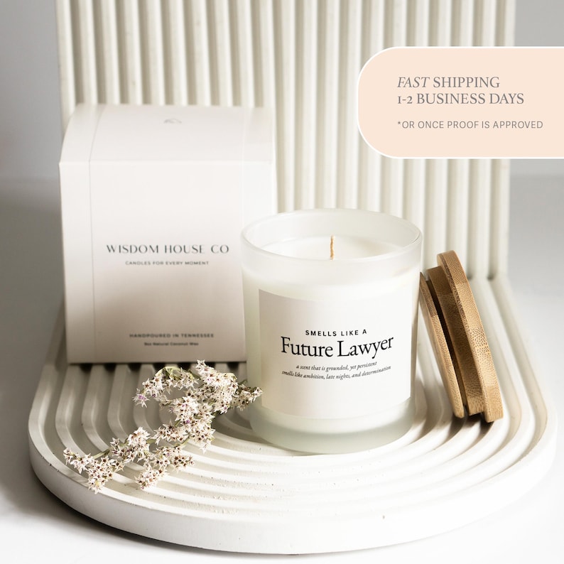 May include: A white candle in a frosted glass jar with a wooden lid. The label reads "Smells Like a Future Lawyer." A white box with the text "Wisdom House Co" is behind the candle. The candle sits on a white, curved tray.